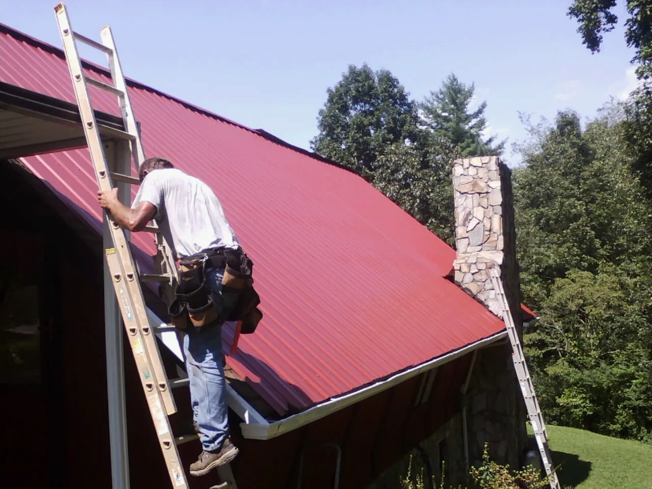 Roofer on a ladder beside a red metal roof and stone chimney — Soffit & Fascia Repair by Roofing Brothers in Newtonsville